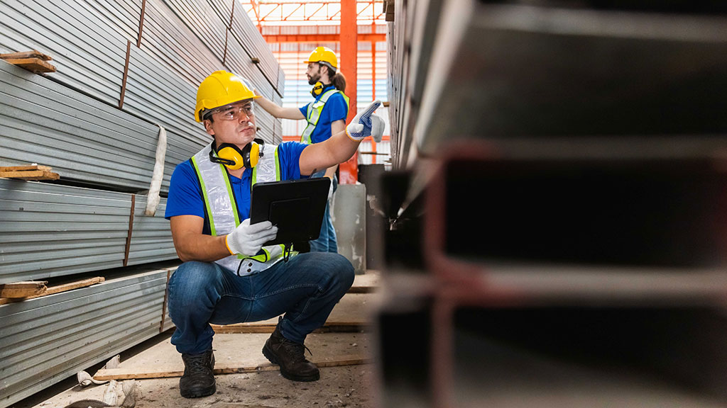 A worker wearing safety gear with a digital tablet is inspecting steel building materials on a rack in a warehouse