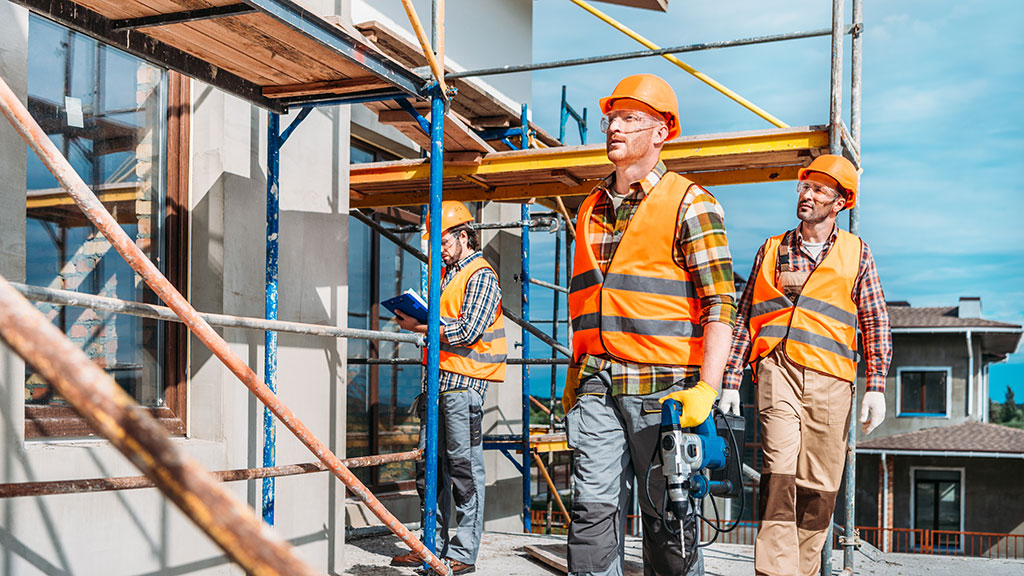 Builders in high-vis vests and hard hats walking beside scaffolding on a residential site, carrying tools.