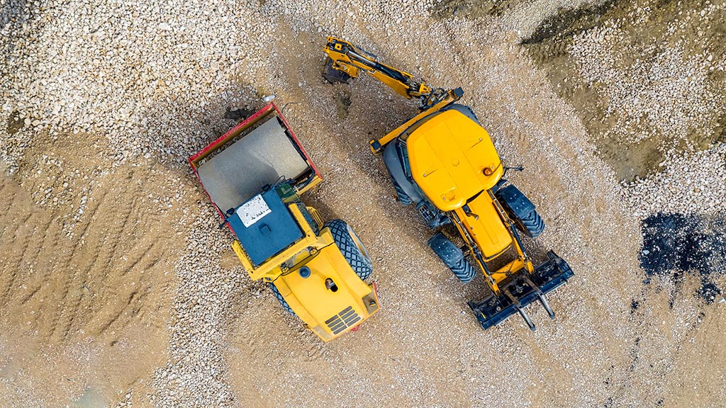 Aerial top-down view of a yellow site dumper beside a yellow backhoe loader with pallet forks parked on gravel at a construction site.