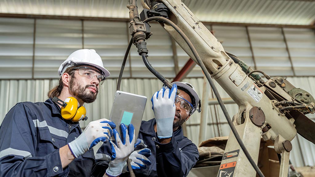 Two engineers in PPE inspect an industrial robotic arm, reviewing diagnostics on a tablet inside a factory workshop.