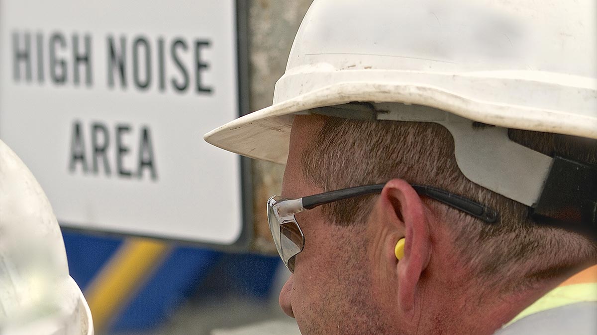Man wearing a hard hat, safety glasses and ear plugs standing near high noise area sign