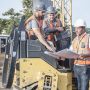 Construction crew in hi-vis vests and hard hats reviewing site plans on a yellow compact roller at a building site with a crane, rebar and shipping containers in view.