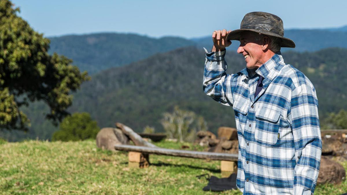 Man wearing blue shirt, smiling and standing in a farming landscape holding his hat
