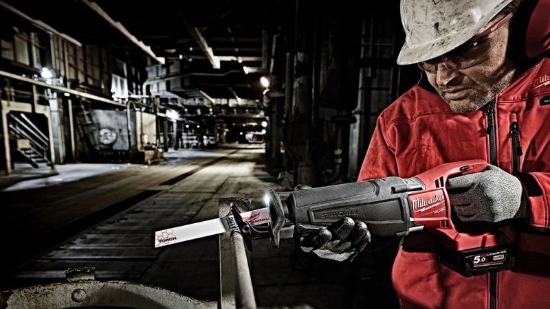 A worker in a darkly lit factory wearing a white hard hard and safety goggles cutting a steel pipe with a Milwaukee reciprocating saw