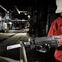 A worker in a darkly lit factory wearing a white hard hard and safety goggles cutting a steel pipe with a Milwaukee reciprocating saw