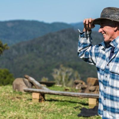 Man wearing blue shirt, smiling and standing in a farming landscape holding his hat